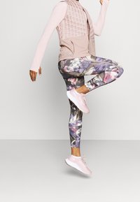 Osoba w różowej bluzce z długim rękawem, jasnoróżowej kamizelce, legginsach w kwiatowy wzór i jasnoróżowych sneakersach, podnosząca jedną nogę w powietrzu na jednolitym tle.