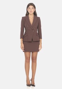 Blazer marrone doppiopetto con bottoni dorati, design aderente e due tasche frontali, abbinato a una gonna mini coordinata. Le scarpe con tacco alto completano il look.