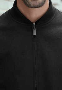 Juodas zip-up džemperis su grotuotu apykakle, pagamintas iš minkštos medžiagos. Savybės: sidabrinis užtrauktuko traukiklis ir lygus paviršius.