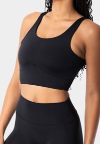 Sujetador deportivo negro con una banda ancha y una textura suave. Presenta un escote en forma de scoop y un detalle de costura en ángulo debajo del busto.