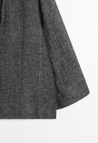 Tissu tricoté gris foncé avec un motif tissé texturé, montrant la manche et une partie du torse sur un fond blanc uni.