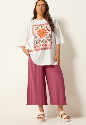 Kobieta ubrana w oversize białą koszulkę z grafiką słońca, księżyca i kwiatów z napisem „Sun Seeker”, zestawioną z luźnymi, różowymi, szerokimi spodniami i białymi sandałami.