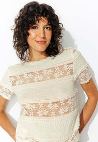 Haut en dentelle crème avec des motifs floraux en dentelle horizontale et une coupe décontractée. Manches courtes avec des accents texturés et un col à bords festonnés.