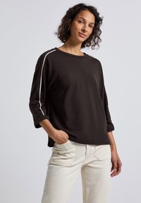 Schwarzes langärmliges Shirt mit weißen Seitenstreifen, Rundhalsausschnitt und lockerer Passform, kombiniert mit hellen Hosen.