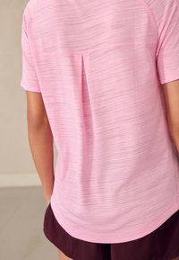 Hellrosa Sportshirt mit strukturiertem Muster, kurzen Ärmeln und Detail mit Schlitz am Rücken, kombiniert mit dunkelbraunen Shorts.