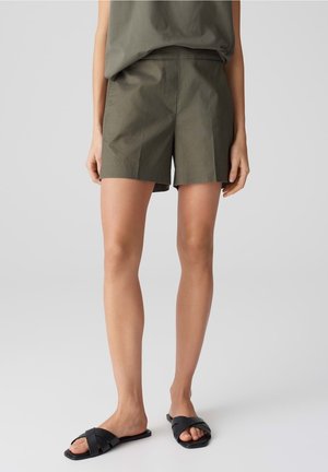Shorts en coton vert olive avec une taille plate, des poches sur les côtés et une coupe décontractée, associés à des sandales noires à glissière avec des lanières entrecroisées.