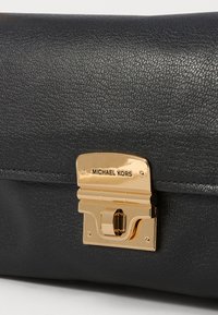 Sort læderhåndtaske med en guldfarvet hardware-lås med "Michael Kors" logo, struktureret overflade og rundet design.
