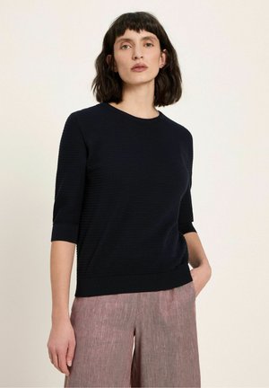 Frau mit kurzem dunklem Haar, die ein schwarzes, geripptes Strickoberteil mit Ellbogen-langen Ärmeln und lockeren, rosa strukturierten Hosen trägt, steht mit einer Hand in der Tasche.