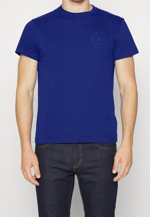 Homme portant un t-shirt bleu roi uni à manches courtes avec un petit logo circulaire sur la poitrine gauche et un jean bleu foncé.
