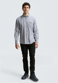 Chemise boutonnée en tissu gris clair avec une texture subtile, dotée d'une poche poitrine, associée à un pantalon noir et des chaussures foncées.
