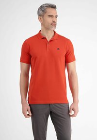 Czerwona koszulka polo z krótkim rękawem, kołnierzykiem i granatowym logo. Materiał wydaje się gładki z delikatną teksturą. Połączona z szarymi spodniami.