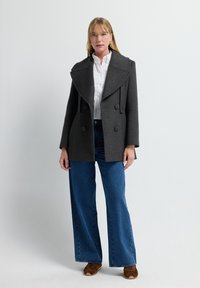 Abrigo de lana gris con grandes solapas y botones de doble botonadura, combinado con jeans de pierna ancha azul y zapatos marrones. Diseño sencillo y estructurado.