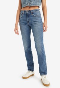 Mujer con jeans rectos de tiro medio en azul, chaleco de denim azul cortado y zapatillas blancas con suelas de goma, de pie frente a un fondo blanco.