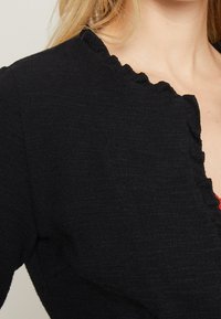 Gros plan sur une personne portant un haut noir texturé avec un col en forme de vague, montrant une partie de l'épaule et du cou.