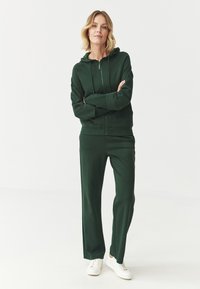 Sudadera con cremallera verde y pantalones de chándal a juego en una tela suave. Presenta una cremallera frontal y una capucha. Combinada con zapatillas blancas.