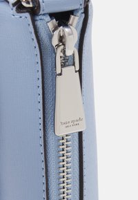 Ljusblå läderväska med dragkedja, utrustad med en silverfärgad metallflärp med ingraverat varumärkesnamn och texturerade materialdetaljer runt dragkedjan.