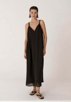 Kobieta w czarnej sukience maxi bez rękawów z podwójnymi cienkimi ramiączkami i czarnych sandałach, stojąca na tle jednolitego jasnego tła.