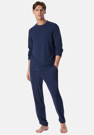 Giovane uomo in piedi a piedi nudi, indossa una maglia blu navy a maniche lunghe e pantaloni abbinati con le mani in tasca su uno sfondo semplice.
