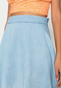 Hellblauer Jeansrock mit glatter Textur, ausgestattet mit einem taillierten Bund und einem ausgestellten Design, teilweise von der Taille abwärts gezeigt.