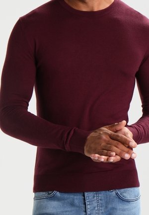 Mann trägt einen burgunderfarbenen Langarmpullover und blaue Jeans, hält die Hände vor dem Oberkörper verschränkt vor einem schlichten hellen Hintergrund.