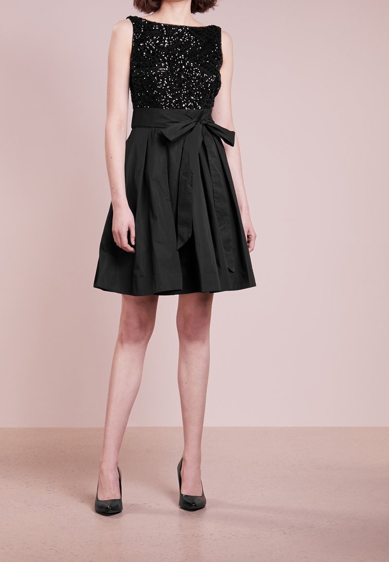 Černé šaty s korzetem zdobeným flitry a rozšířenou sukní, s páskem v oblasti pasu. Šaty mají jemnou texturu a jsou do délky po kolena.