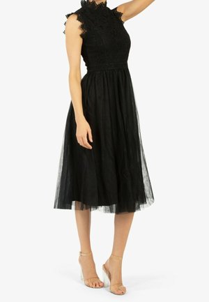 Woman wearing a sleeveless black lace midi dress with a high neck and sheer overlay, paired with clear strap high-heeled sandals.