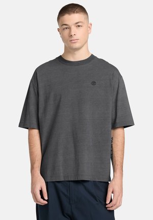 Giovane uomo con capelli corti che indossa una maglietta grigio scuro ampia con un piccolo logo nero sul petto a sinistra e pantaloni scuri.