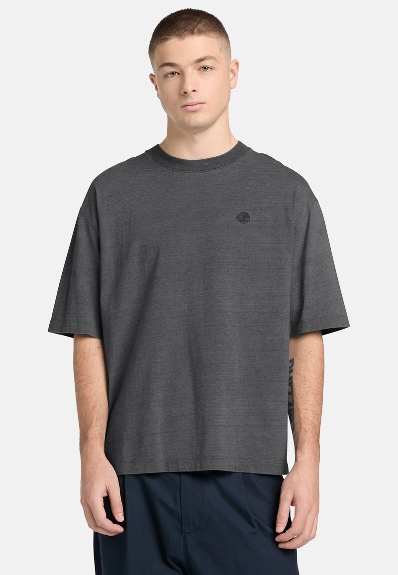 Jeune homme aux cheveux courts portant un t-shirt gris foncé ample avec un petit logo noir sur la poitrine gauche et un pantalon foncé.