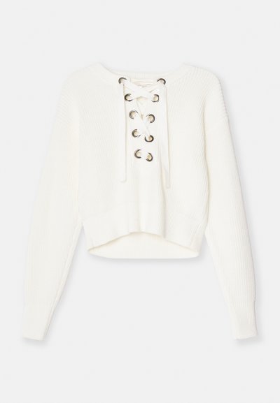 Fehér, rövidített pulóver, bordás kötésű anyagból készült. Fűzős eleje fém szemekkel díszített. Hosszú ujjú és széles, bordás szegéllyel rendelkezik.