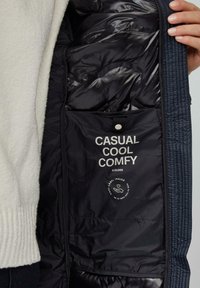 Quiltana mornarsko modra jakna z notranjostjo z mehko črno podlogo, označena z "CASUAL COOL COMFY" v belem, vključuje žep z gumbi za shranjevanje.