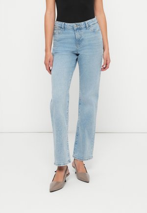 Kobieta w jasnoniebieskich jeansach o prostych nogawkach i taupe szpilkach z odkrytymi piętami, stojąca na białym tle.