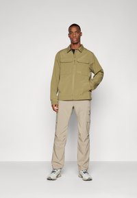 Veste zippée vert olive avec deux poches avant, associée à un pantalon cargo beige clair doté de poches latérales et à des baskets grises.