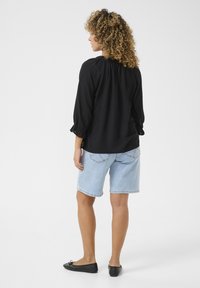 Schwarze Bluse mit langen Ärmeln und gerafftem Ausschnitt, kombiniert mit hellblauen Jeansshorts. Dazu flache schwarze Ballerinas. Schlichter Hintergrund.