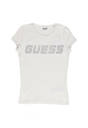 Bijela majica prilagođena tijelu izrađena od pamuka s kratkim rukavima, s istaknutim logom "GUESS" u srebrnim ukrasima. Glatka tekstura.