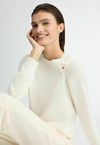 Pull en maille couleur crème avec des accents côtelés aux épaules et un détail de bouton sur un côté. Texture lisse et coupe décontractée.