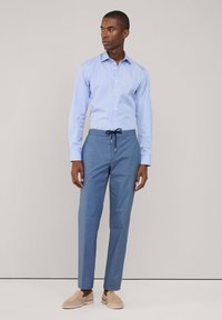 Lichtblauwe gestreepte blouse met een klassieke kraag, gecombineerd met effen blauwe broeken met een trekkoord in de taille. Het schoeisel is lichtbeige.