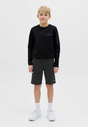 Felpa nera con scollatura rotonda, abbinata a pantaloni corti grigio antracite. Presenta il logo sulla felpa e sneakers bianche con calzini alla caviglia.