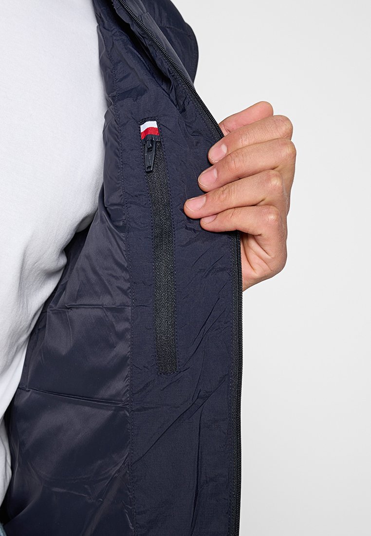 Chaleco de plumón azul marino con un bolsillo interior con cremallera. Presenta un pequeño acento en rojo, blanco y azul cerca del bolsillo. Tejido texturizado con un acabado arrugado.