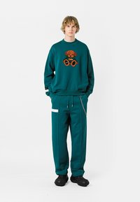 Sudadera teal con bordado de oso de peluche, combinada con pantalones jogger teal a juego que tienen bolsillos laterales y detalle de cremallera, usados con zapatos negros.