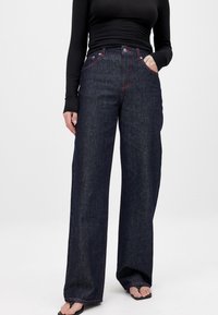 Temno modri široki džins hlače iz denima, s visokim pasom, rdečimi šivi in sprednjimi žepi. Nosijo se z črnim dolgem rokavom.