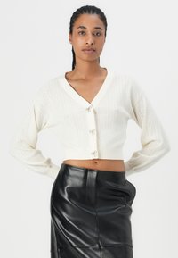 Gilet court crème avec un décolleté en V, motif tricoté texturé et fermeture à trois boutons. Associé à une jupe en cuir noir taille haute.