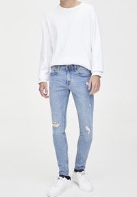 Jeans ajustados de color azul claro con parches desgastados, combinados con una camisa blanca de manga larga y zapatillas blancas, de pie frente a un fondo liso.