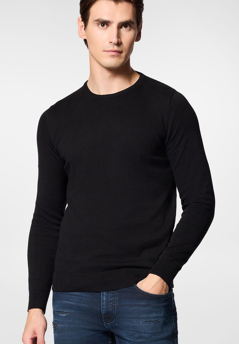 Czarny dzianinowy sweter z długimi rękawami i okrągłym dekoltem. Posiada ściągacze na mankietach i u dołu. Noszony z niebieskimi dżinsami.