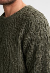 Olive'owa zielona sweter z dzianiny o splocie kablowym z okrągłym dekoltem, teksturowanymi wzorami i grubym wykończeniem wełnianym. Brak widocznych elementów metalowych lub akcentów.