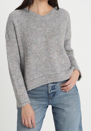 Femme portant un pull en maille côtelée gris à manches longues et un jean taille haute bleu clair, main reposant sur la poche du jean.