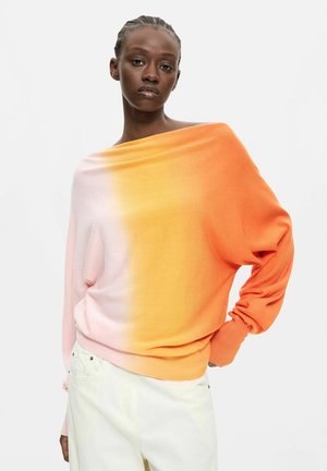 Ung kvinde med fletninger iført en gradient off-shoulder sweater, der går fra pink til orange, og hvide bukser mod hvid baggrund.