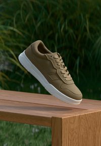 Ruskeat lenkkarit, joissa on röpelöinen pinta, valkoinen pohja ja perinteinen nauhakiinnitys, esitettynä puupinnalla luonnollista taustaa vasten.