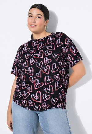 Kobieta o ciemnych włosach, ubrana w czarny t-shirt pokryty różowymi i jasnoniebieskimi wzorami serc, w połączeniu z jasnoniebieskimi dżinsami.