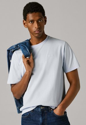 Mladić u svijetloplavoj majici i tamnim trapericama, drži plavu traper jaknu preko jednog ramena na jednobojnoj sivoj pozadini.