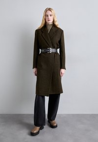 Oliwkowy wełniany płaszcz o sztywnym fasonie, ściśnięty szerokim czarnym paskiem z srebrnymi detalami, w połączeniu z czarnymi spodniami o szerokich nogawkach i brązowymi botkami.